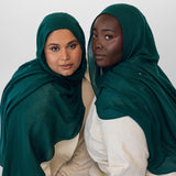 Two women wearing evergreen hijabs and white clothing against a white background