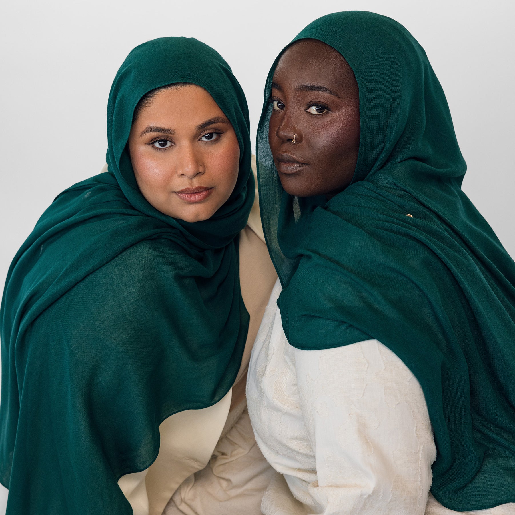 Two women wearing evergreen hijabs and white clothing against a white background
