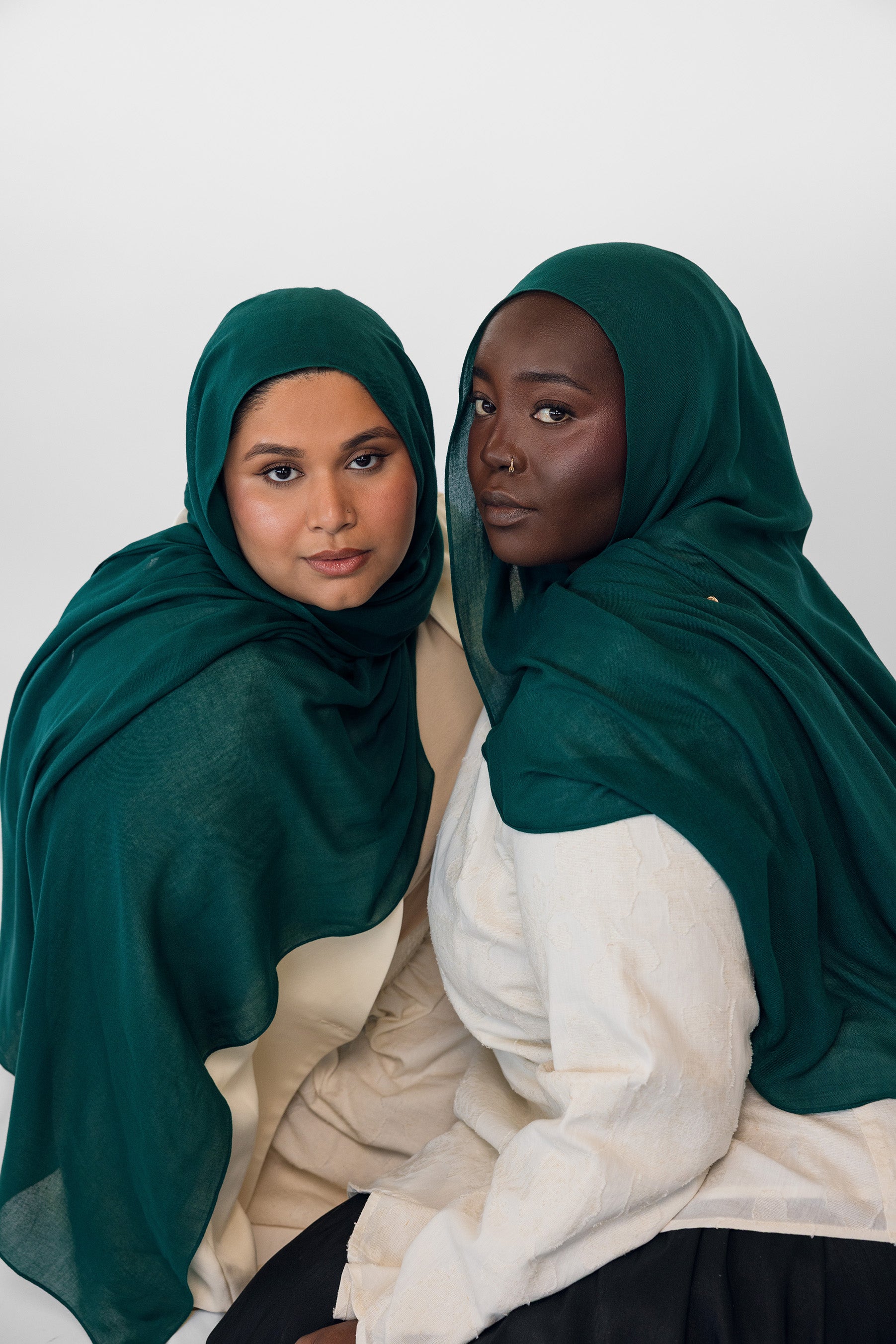 Two women wearing evergreen hijabs and white clothing against a white background