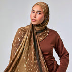 A woman wearing a brown and beige patterned hijab draped over her shoulders and a rust-brown long-sleeve top, photographed against a plain light gray studio background.