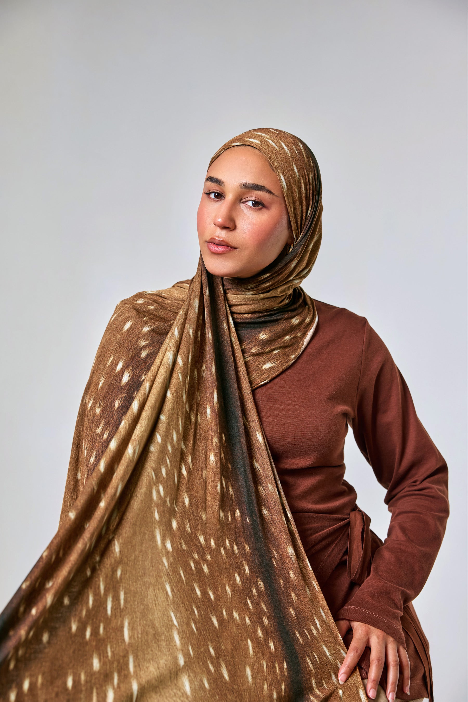 A woman wearing a brown and beige patterned hijab draped over her shoulders and a rust-brown long-sleeve top, photographed against a plain light gray studio background.