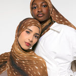 Two models wearing brown and beige spotted hijabs, with one in a white shirt, photographed against a clean white studio background.