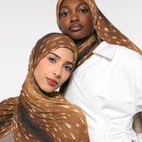 Two models wearing brown and beige spotted hijabs, with one in a white shirt, photographed against a clean white studio background.