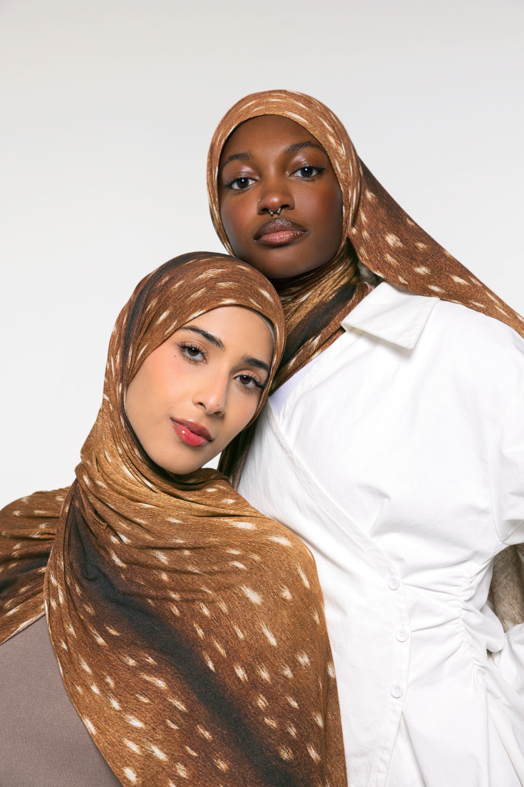Two models wearing brown and beige spotted hijabs, with one in a white shirt, photographed against a clean white studio background.