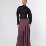 A model stands against a light gray studio background wearing a black long-sleeve mock-neck top and a high-waisted, pleated mauve-brown maxi skirt.