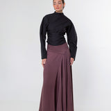 A model stands against a light gray studio background wearing a black long-sleeve mock-neck top and a high-waisted, pleated mauve-brown maxi skirt.