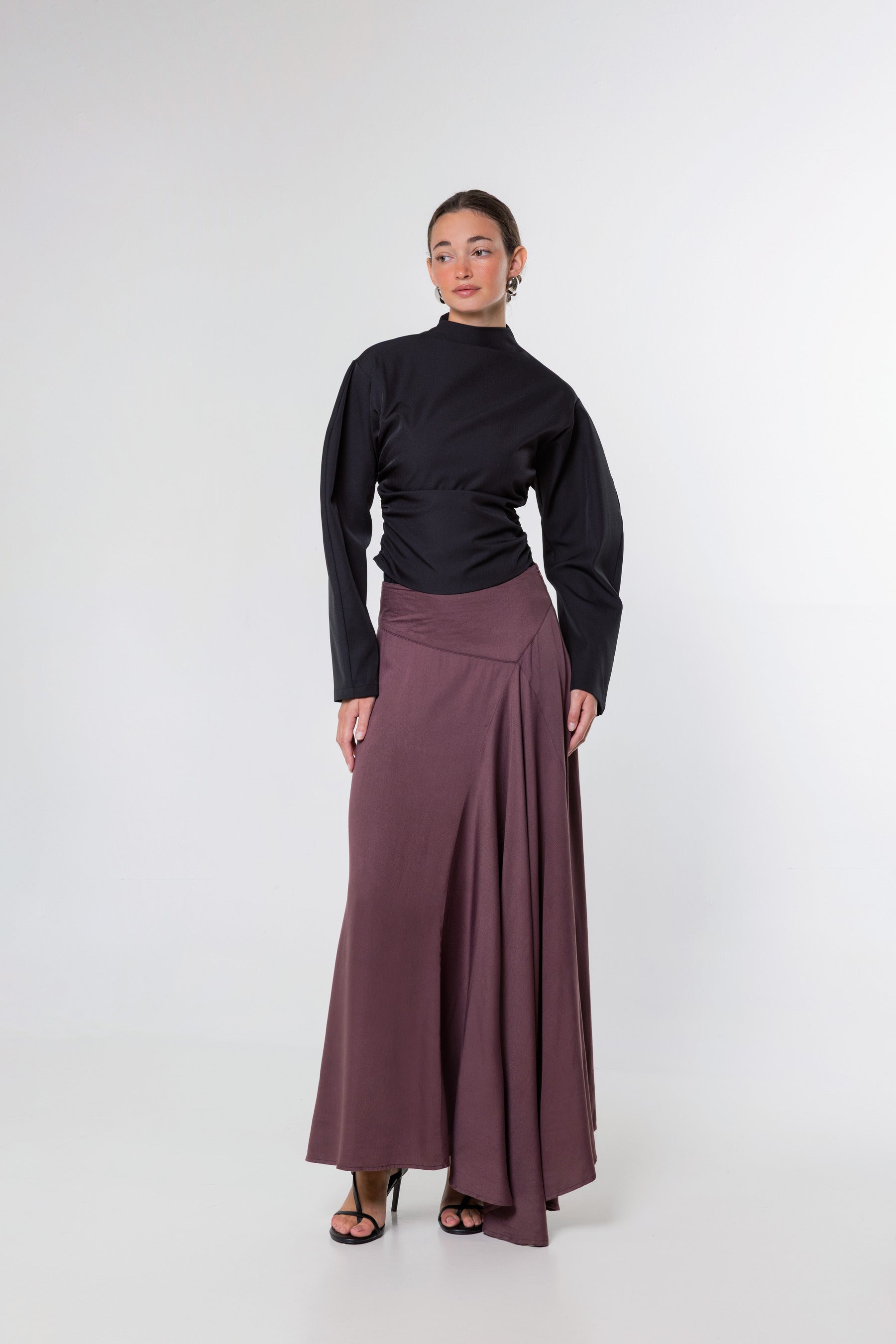 A model stands against a light gray studio background wearing a black long-sleeve mock-neck top and a high-waisted, pleated mauve-brown maxi skirt.