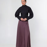 A model stands against a light gray studio background wearing a black long-sleeve mock-neck top and a long, flowing mauve-brown skirt.