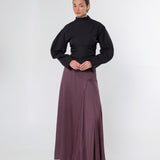 A model stands against a light gray studio background wearing a black long-sleeve mock-neck top and a long, flowing mauve-brown skirt.