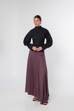 A model stands against a light gray studio background wearing a black long-sleeve mock-neck top and a long, flowing mauve-brown skirt.