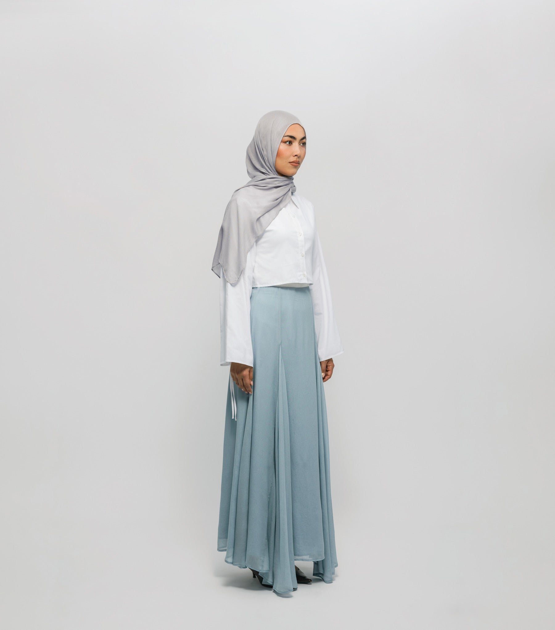 Woman wearing a light gray hijab, white top, and light blue skirt on a white background