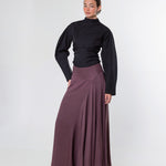 A woman modeling a black long-sleeve mock-neck top and a high-waisted, pleated mauve-brown maxi skirt against a plain light gray studio background.