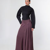 A woman modeling a black long-sleeve mock-neck top and a high-waisted, pleated mauve-brown maxi skirt against a plain light gray studio background.