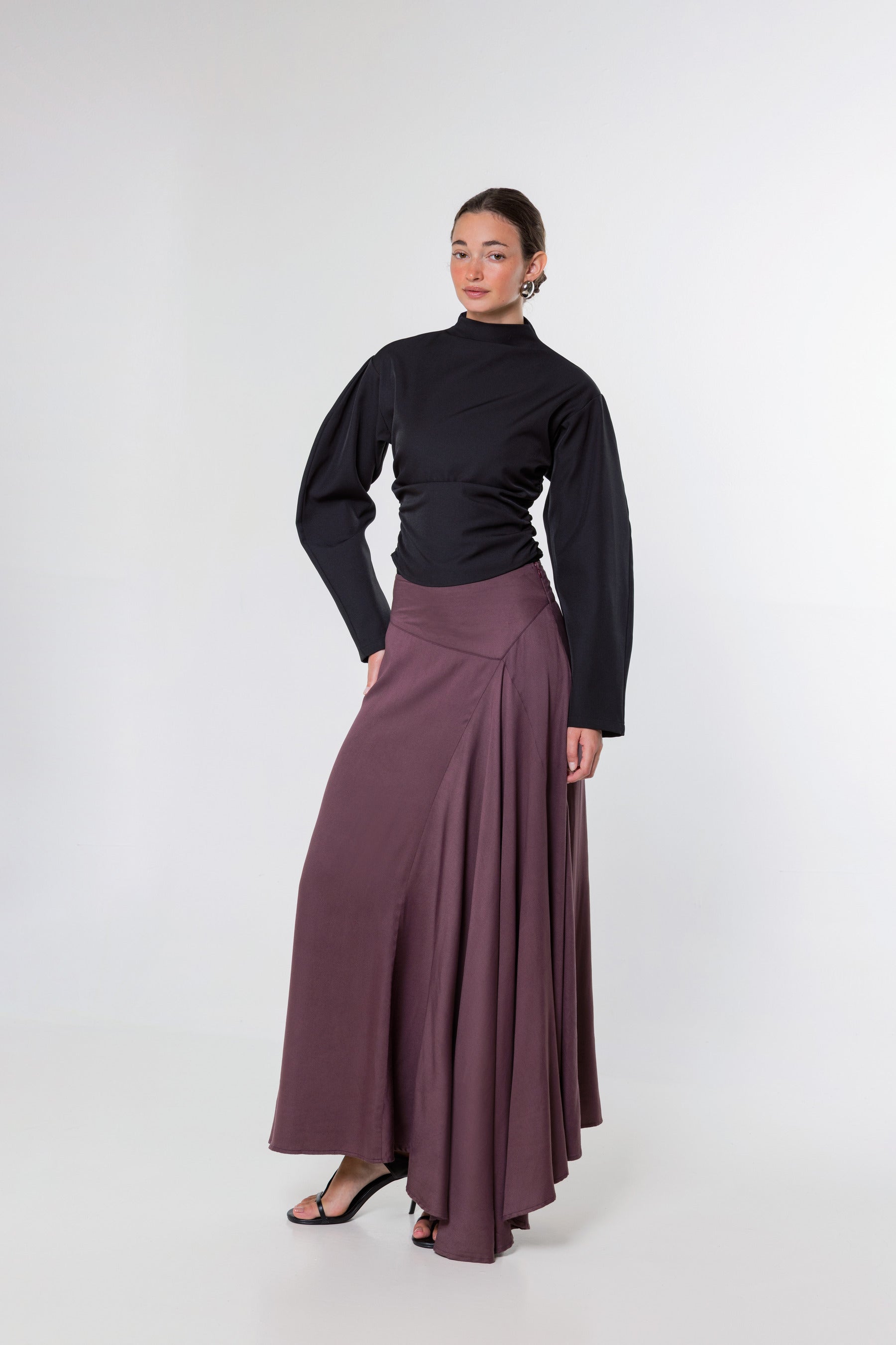 A woman modeling a black long-sleeve mock-neck top and a high-waisted, pleated mauve-brown maxi skirt against a plain light gray studio background.
