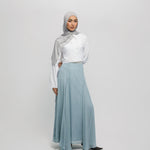 Woman wearing a hijab, white blouse, and light blue skirt on a plain background