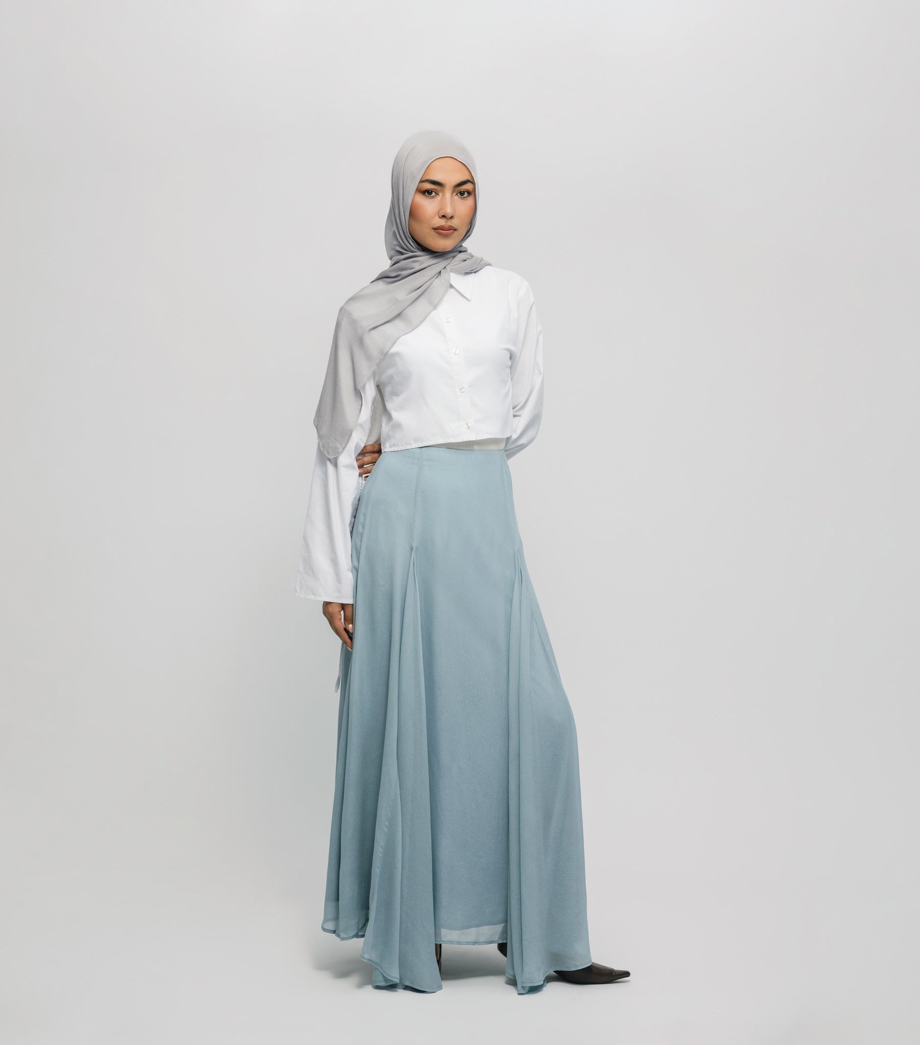 Woman wearing a hijab, white blouse, and light blue skirt on a plain background