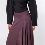 A model shown from the back wearing a black long-sleeve top and a high-waisted, pleated mauve-brown skirt against a light gray studio background.