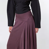 A model shown from the back wearing a black long-sleeve top and a high-waisted, pleated mauve-brown skirt against a light gray studio background.