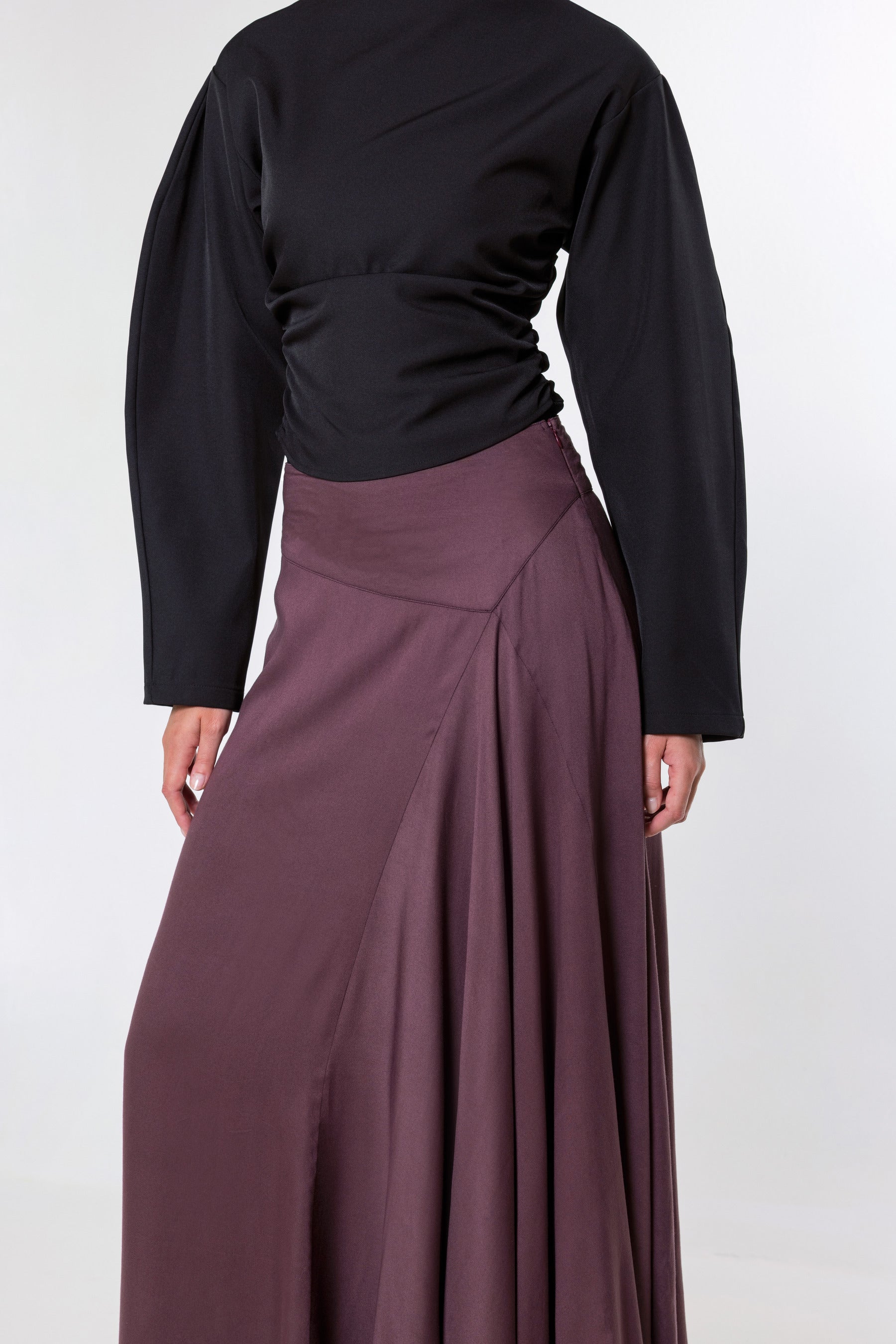 A model shown from the back wearing a black long-sleeve top and a high-waisted, pleated mauve-brown skirt against a light gray studio background.