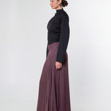 A woman in side profile wearing a black long-sleeve mock-neck top and a high-waisted, pleated mauve-brown maxi skirt, standing against a plain light gray studio background.