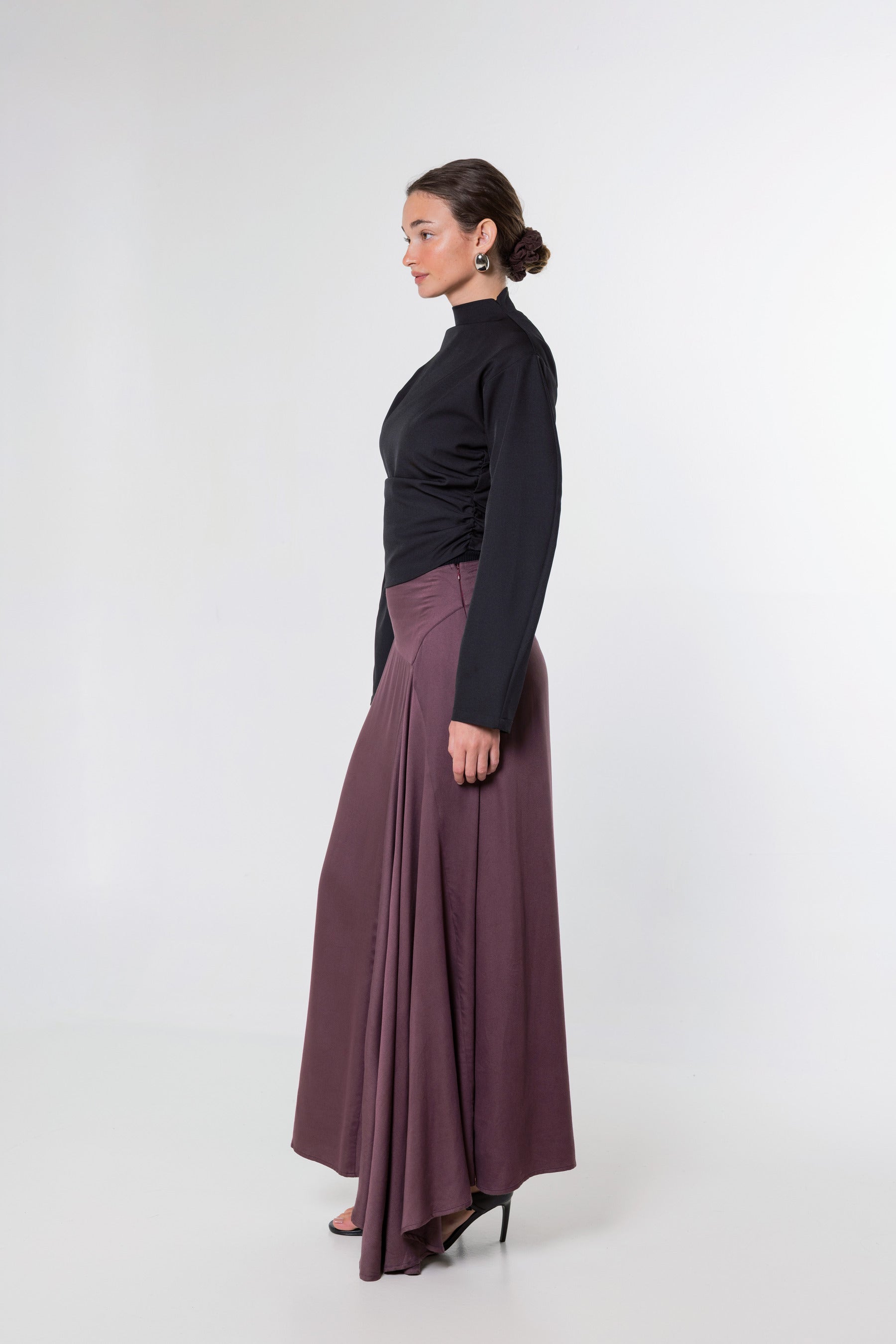 A woman in side profile wearing a black long-sleeve mock-neck top and a high-waisted, pleated mauve-brown maxi skirt, standing against a plain light gray studio background.