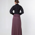 A woman shown from the back wearing a black long-sleeve top and a long mauve-purple skirt, standing against a plain light gray studio background.