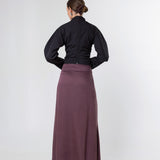 A woman shown from the back wearing a black long-sleeve top and a long mauve-purple skirt, standing against a plain light gray studio background.
