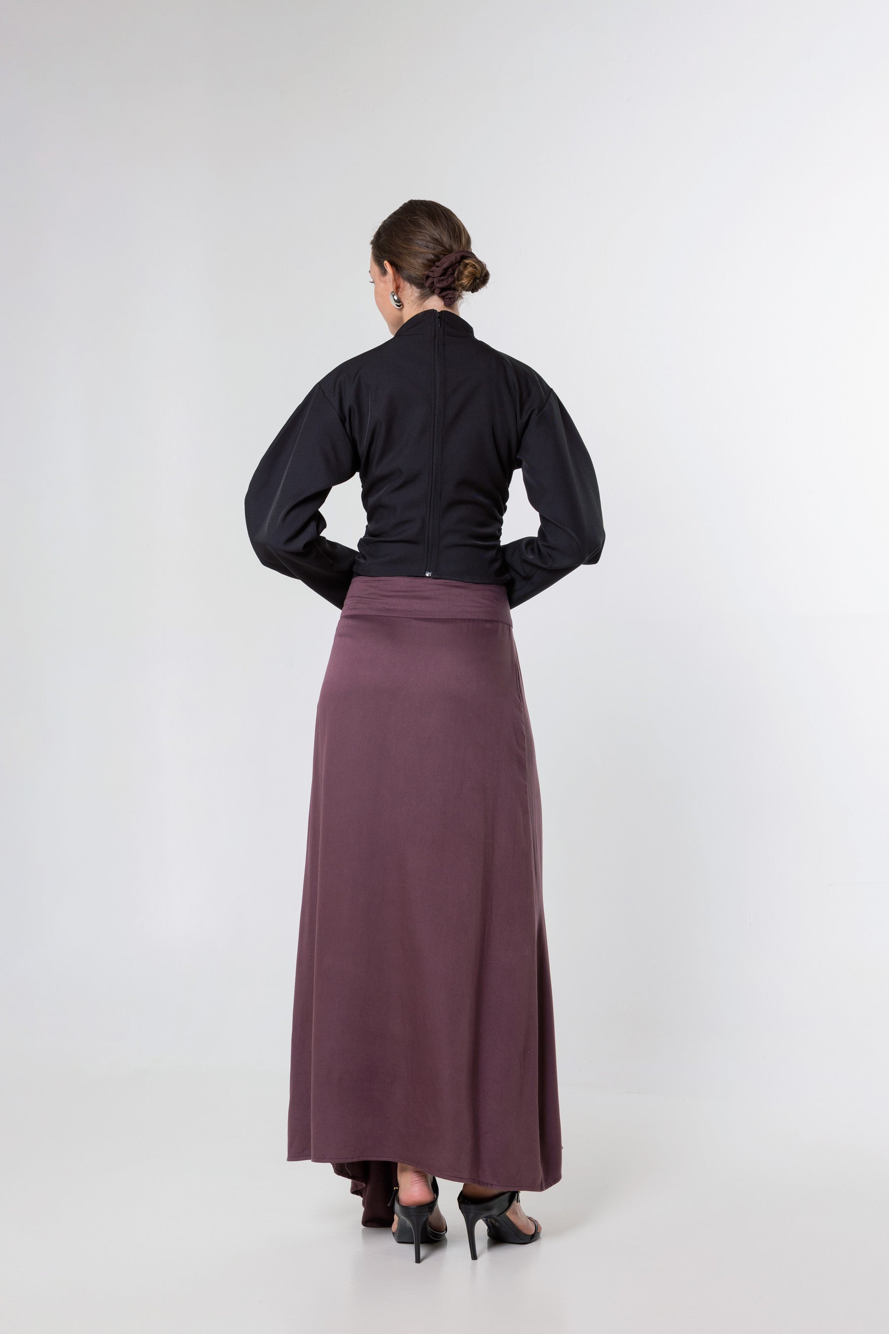 A woman shown from the back wearing a black long-sleeve top and a long mauve-purple skirt, standing against a plain light gray studio background.