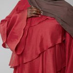Person wearing a red layered dress and brown hijab against a plain background