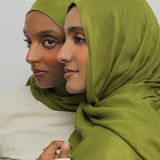 Two women wearing green hijabs and white clothing against a plain background