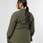 A woman shown from the back wearing an olive green long-sleeve wrap-style top and black pants, standing against a plain light gray studio background.