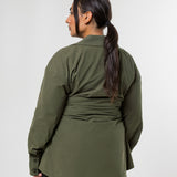 A woman shown from the back wearing an olive green long-sleeve wrap-style top and black pants, standing against a plain light gray studio background.