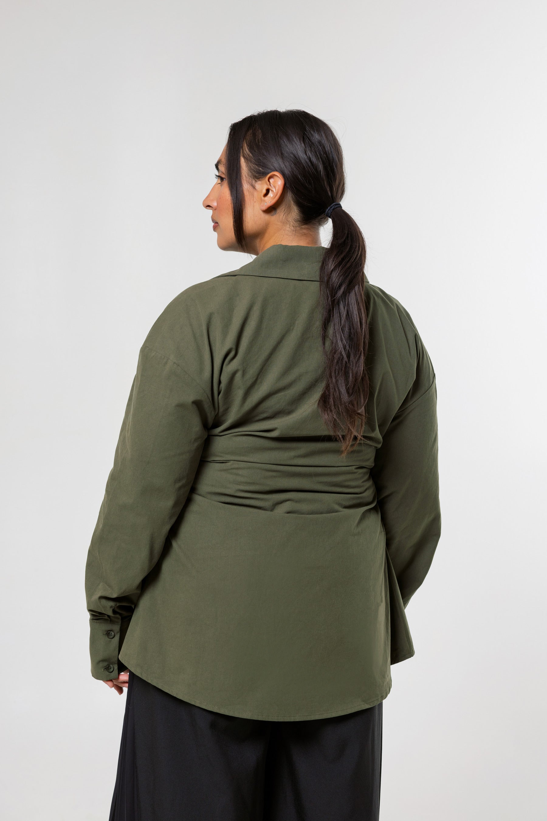 A woman shown from the back wearing an olive green long-sleeve wrap-style top and black pants, standing against a plain light gray studio background.