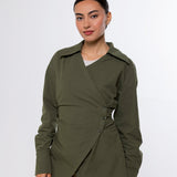 A woman modeling an olive green wrap-style long-sleeve shirt jacket with a buttoned waist detail, paired with beige pants, against a plain light gray studio background.