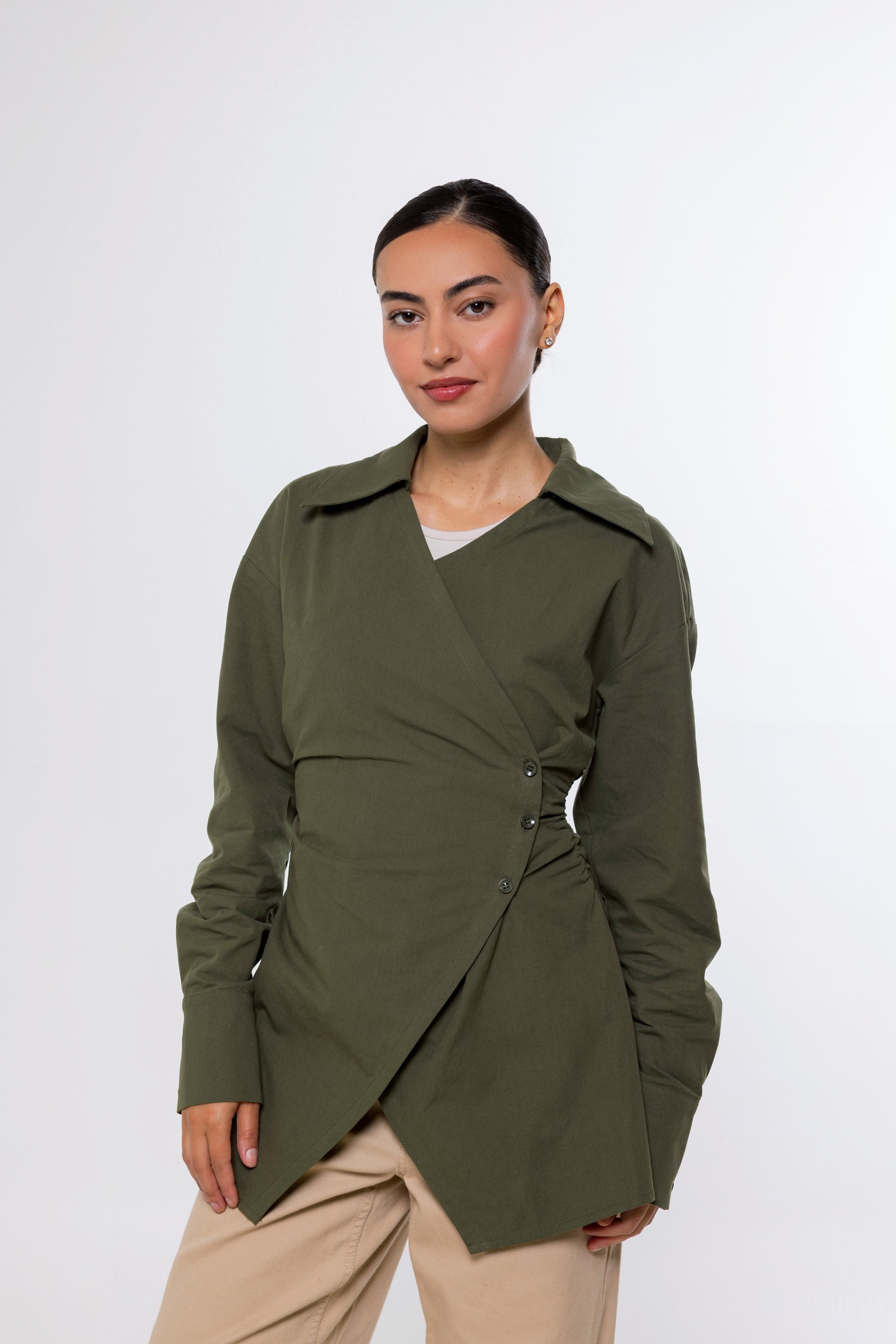 A woman modeling an olive green wrap-style long-sleeve shirt jacket with a buttoned waist detail, paired with beige pants, against a plain light gray studio background.