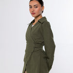 A woman modeling an olive green wrap-style long-sleeve shirt with a gathered waist detail, paired with beige pants, against a plain light gray studio background.