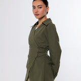 A woman modeling an olive green wrap-style long-sleeve shirt with a gathered waist detail, paired with beige pants, against a plain light gray studio background.