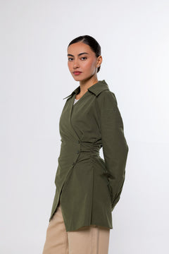 A woman modeling an olive green wrap-style long-sleeve shirt with a gathered waist detail, paired with beige pants, against a plain light gray studio background.