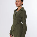 A woman modeling an olive green wrap-style long-sleeve shirt jacket with a cinched waist, paired with beige trousers, against a plain light gray studio background.