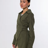 A woman modeling an olive green wrap-style long-sleeve shirt jacket with a cinched waist, paired with beige trousers, against a plain light gray studio background.