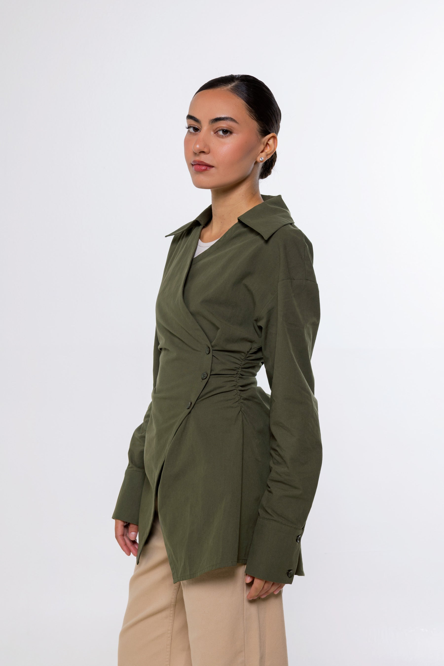 A woman modeling an olive green wrap-style long-sleeve shirt jacket with a cinched waist, paired with beige trousers, against a plain light gray studio background.
