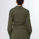 A woman viewed from the back wearing an olive green long-sleeve shirt with a gathered waist detail, paired with beige pants, against a plain light gray background.