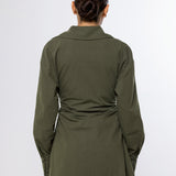A woman viewed from the back wearing an olive green long-sleeve shirt with a gathered waist detail, paired with beige pants, against a plain light gray background.