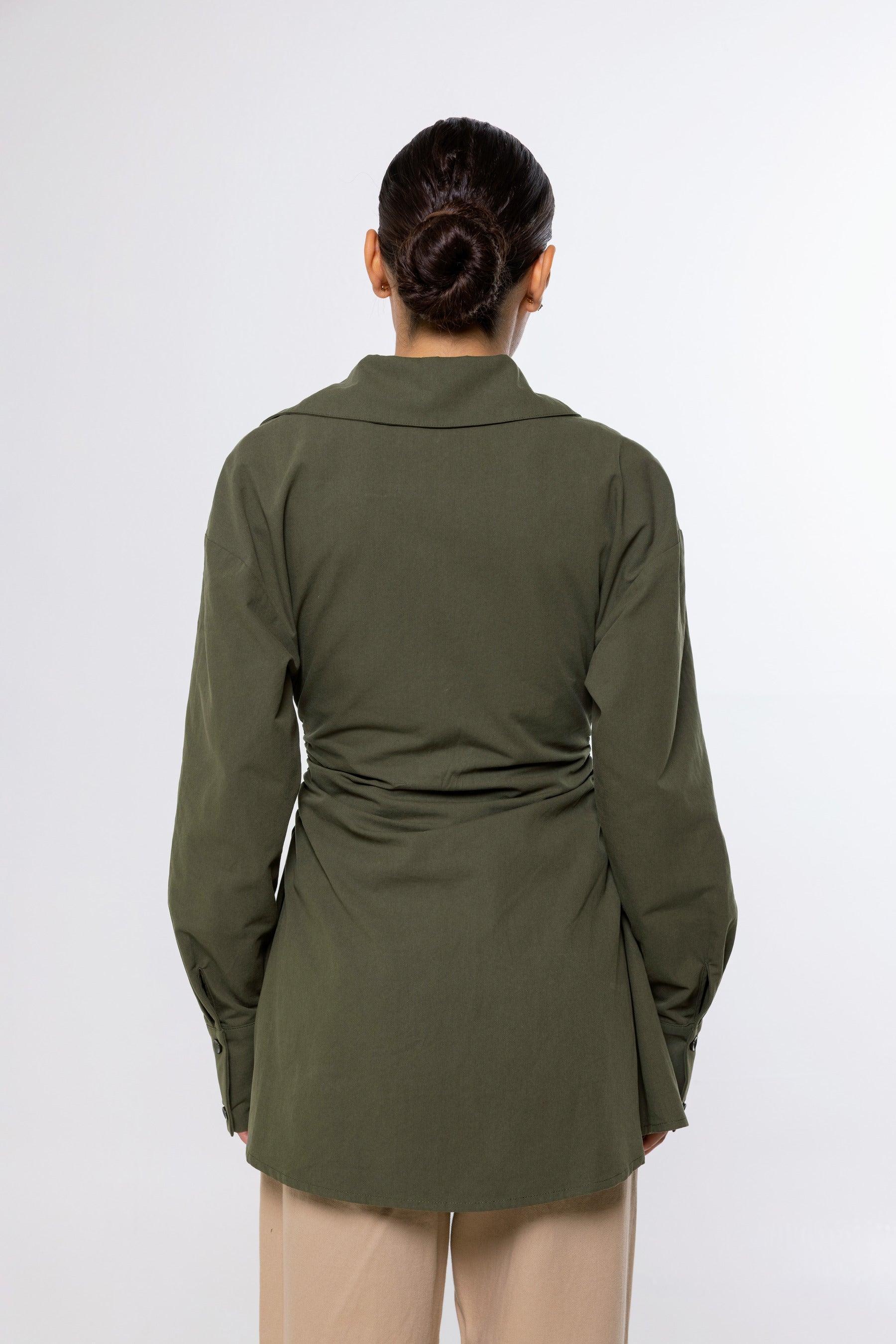 A woman viewed from the back wearing an olive green long-sleeve shirt with a gathered waist detail, paired with beige pants, against a plain light gray background.