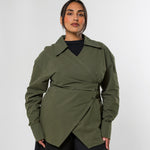 A woman modeling an olive green wrap-style long-sleeve shirt with a belted waist, worn over a black top and black wide-leg pants, against a light gray studio background.