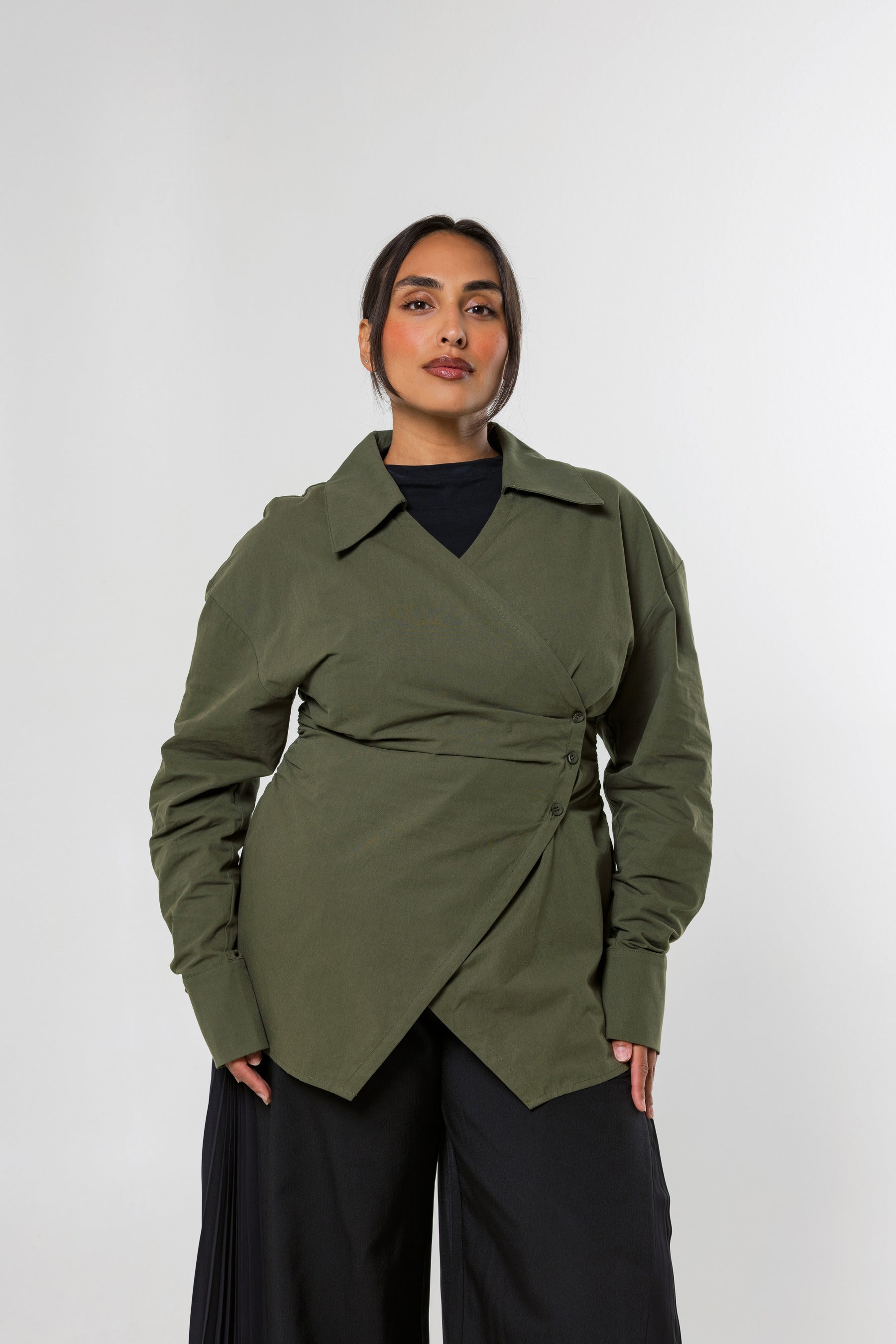 A woman modeling an olive green wrap-style long-sleeve shirt with a belted waist, worn over a black top and black wide-leg pants, against a light gray studio background.