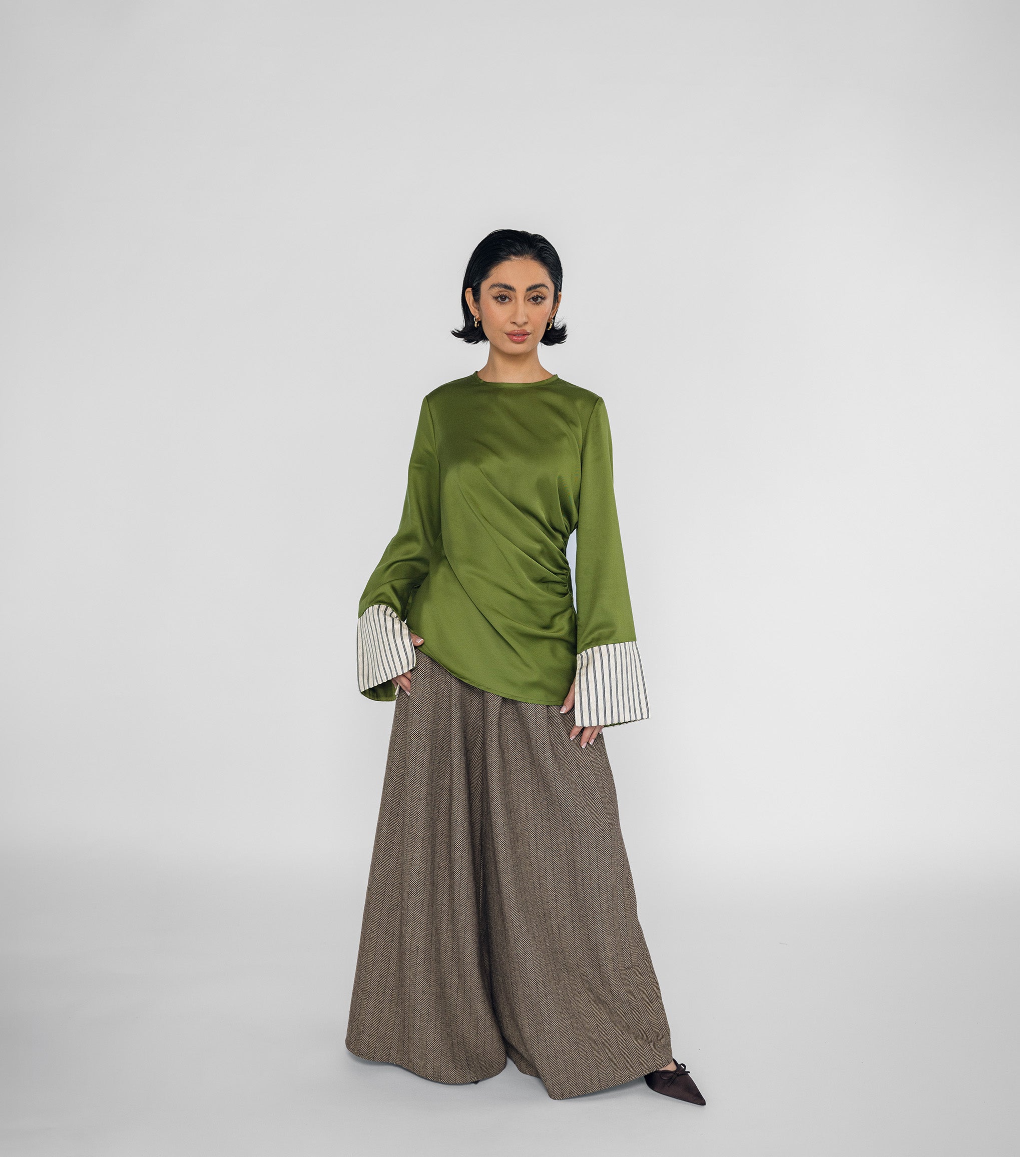Person wearing a green top with white cuffs and brown wide-leg pants on a white background