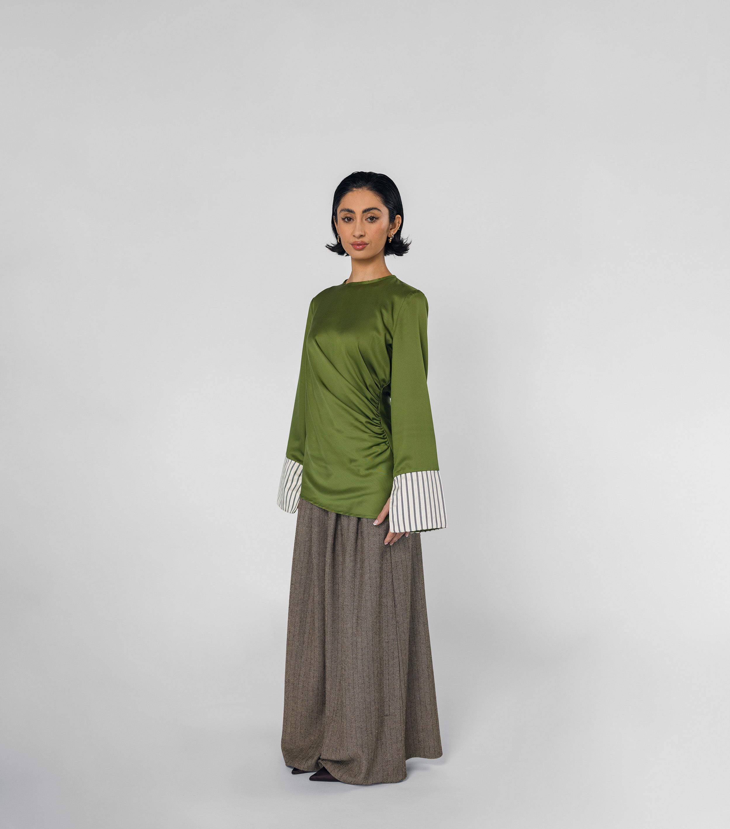Woman wearing a green top with striped cuffs and brown pants on a white background