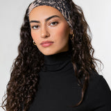 A woman with long, curly dark brown hair is shown in profile wearing a textured lace printed headband with a black turtleneck top against a plain light gray background.
