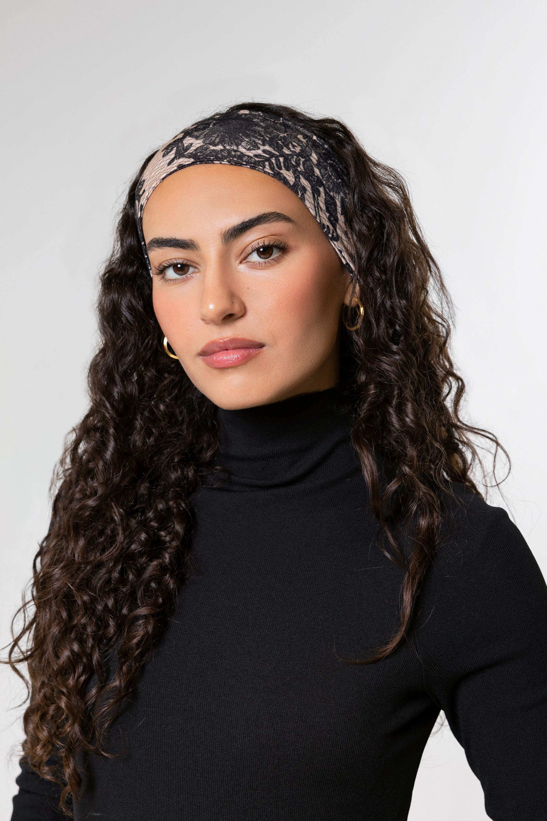 A woman with long, curly dark brown hair is shown in profile wearing a textured lace printed headband with a black turtleneck top against a plain light gray background.